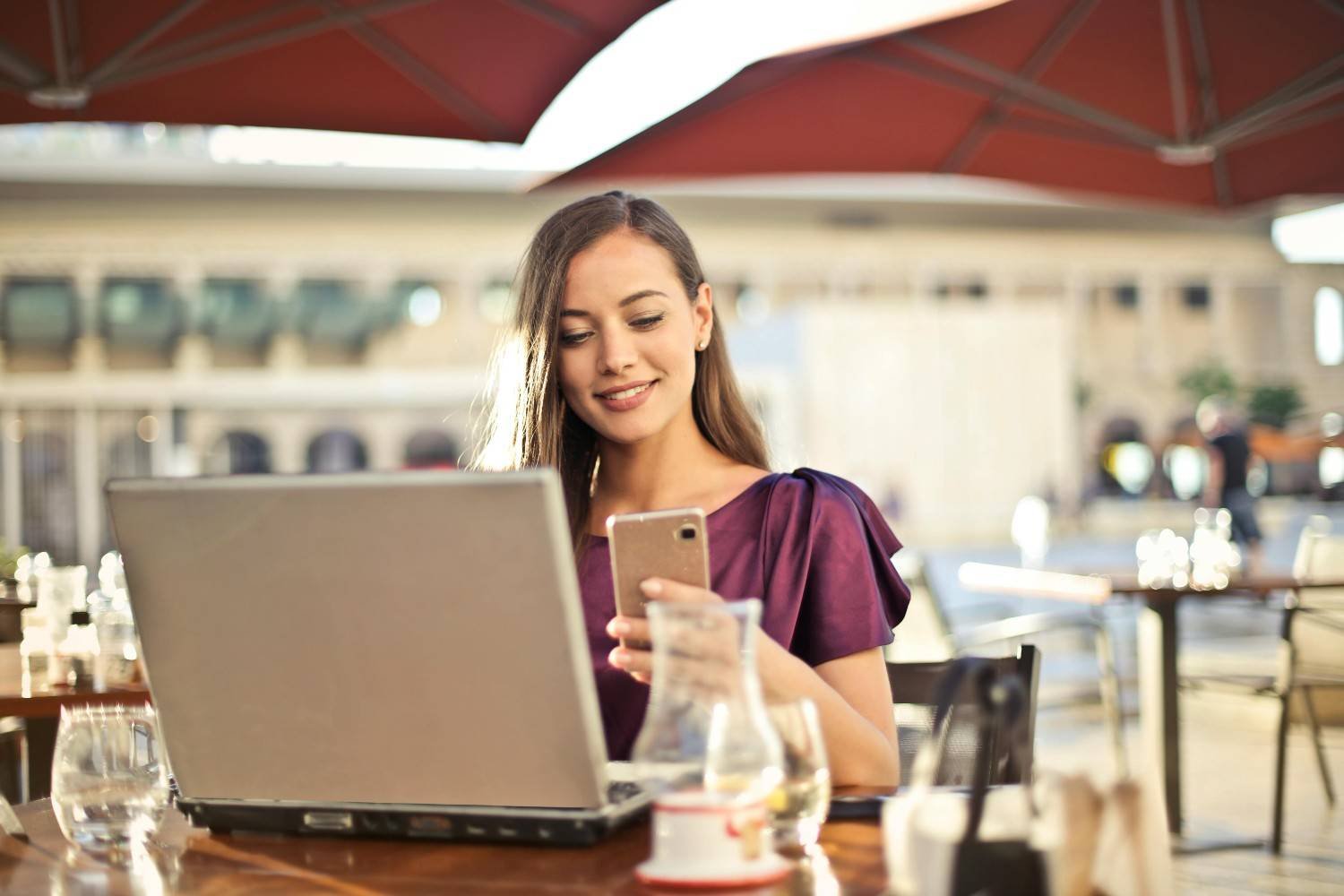 A woman holding her cellphone in front of her laptop while working outdoors
